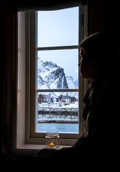 Nachdenkliche Frau schaut aus dem Fenster auf einen verschneiten Berg, Reine, Lofoten, Norwegen - CAIF22620