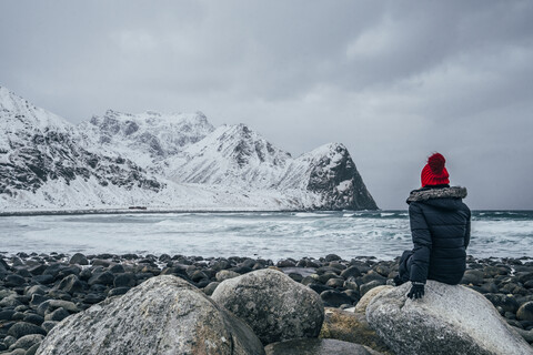 Frau in warmer Kleidung genießt den Blick auf das verschneite Meer und die Berge, Lofoten-Inseln, Norwegen, lizenzfreies Stockfoto