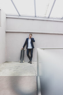 Lächelnder junger Geschäftsmann mit Skateboard, der an einer Betonwand lehnt und mit einem Handy telefoniert - MOEF02080