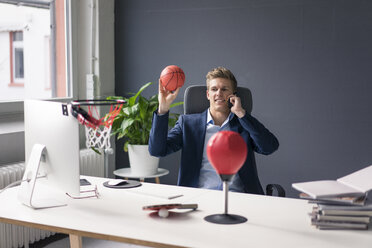 Junger Geschäftsmann sitzt am Schreibtisch im Büro, spielt Basketball und spricht mit seinem Handy - MOEF02081