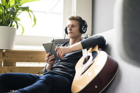 Entspannter junger Mann mit Tablet und Gitarre, der mit Kopfhörern Musik hört - MOEF02082