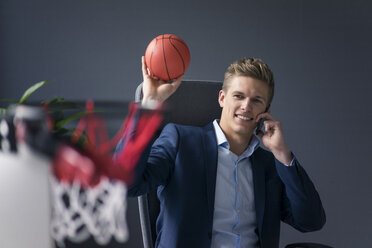 Junger Geschäftsmann sitzt am Schreibtisch im Büro, spielt Basketball und spricht mit seinem Handy - MOEF02085