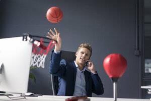 Junger Geschäftsmann sitzt am Schreibtisch im Büro, spielt Basketball und spricht mit seinem Handy - MOEF02089