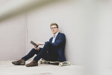 Young businessman with skateboard sitting outdoors using tablet - MOEF02093