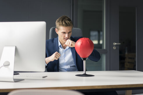 Junger Geschäftsmann am Schreibtisch im Büro beim Boxen mit einem Punchingball - MOEF02094