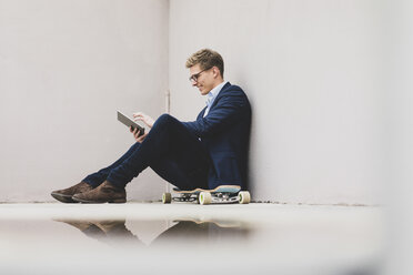 Smiling young businessman with skateboard sitting outdoors using tablet - MOEF02095