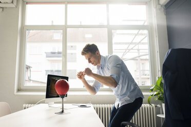 Young businessman at desk in office boxing with punching ball - MOEF02098