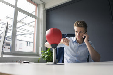 Junger Geschäftsmann am Schreibtisch im Büro, der mit einem Punchingball boxt und mit einem Handy spricht - MOEF02100