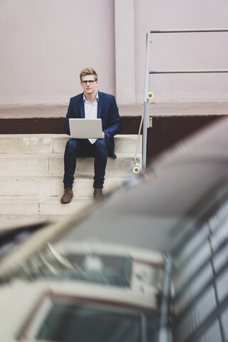 Young businessman with skateboard sitting outdoors on stairs using laptop stock photo