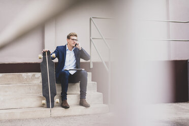 Young businessman with skateboard sitting outdoors on stairs talking on cell phone - MOEF02105