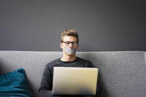 Junger Mann mit zugeklebtem Mund sitzt auf einer Couch und benutzt einen Laptop - MOEF02108