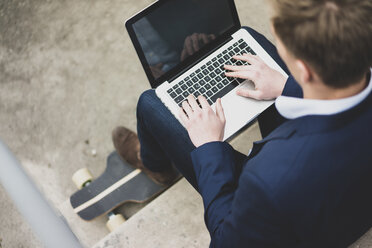 Young businessman with skateboard sitting outdoors on stairs using laptop - MOEF02109