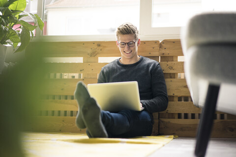 Lächelnder junger Mann, der auf dem Boden sitzt und einen Laptop benutzt, lizenzfreies Stockfoto