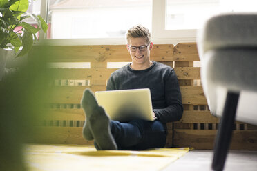 Lächelnder junger Mann, der auf dem Boden sitzt und einen Laptop benutzt - MOEF02112
