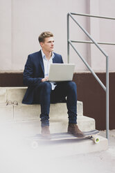 Young businessman with skateboard sitting outdoors on stairs using laptop - MOEF02113