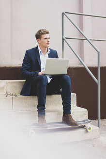 Junger Geschäftsmann mit Skateboard sitzt im Freien auf einer Treppe und benutzt einen Laptop - MOEF02113