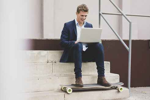 Glücklicher junger Geschäftsmann mit Skateboard, der im Freien auf einer Treppe sitzt und einen Laptop benutzt - MOEF02114
