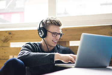 Lächelnder junger Mann, der einen Laptop benutzt und mit Kopfhörern Musik hört - MOEF02115