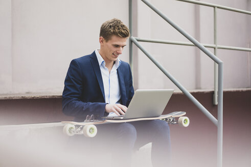 Lächelnder junger Geschäftsmann mit Skateboard, der im Freien auf einer Treppe sitzt und einen Laptop benutzt - MOEF02117