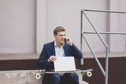 Junger Geschäftsmann mit Skateboard und Laptop sitzt im Freien auf einer Treppe und spricht mit seinem Handy - MOEF02121