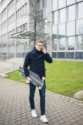 Lächelnder junger Mann mit Skateboard im Gespräch mit dem Handy in der Stadt - MOEF02124