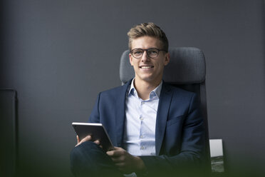 Lächelnder junger Geschäftsmann sitzt auf einem Bürostuhl und benutzt ein Tablet - MOEF02132