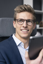 Lächelnder junger Geschäftsmann sitzt auf einem Bürostuhl und benutzt ein Tablet - MOEF02134
