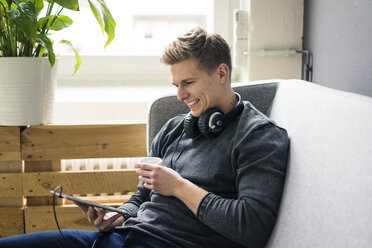Lächelnder junger Mann mit Tablet und Kopfhörern auf der Couch sitzend - MOEF02135