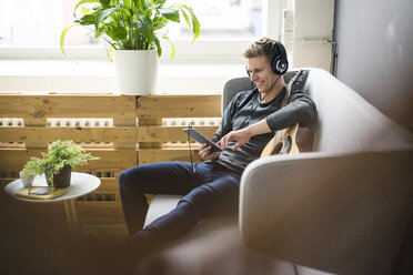 Lächelnder junger Mann mit Gitarre, Tablet und Kopfhörern auf der Couch sitzend - MOEF02138