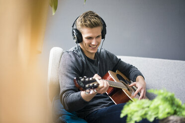 Lächelnder junger Mann mit Kopfhörern, der auf einer Couch sitzt und Gitarre spielt - MOEF02142