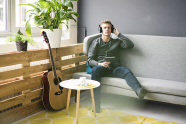 Entspannter junger Mann mit Tablet und Gitarre, der mit Kopfhörern Musik hört - MOEF02143