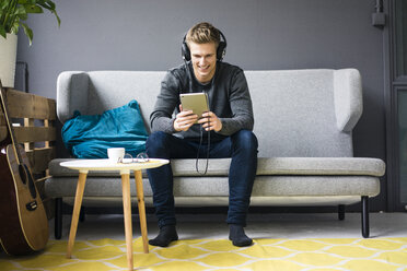 Lächelnder junger Mann mit Gitarre, Tablet und Kopfhörern auf der Couch sitzend - MOEF02145