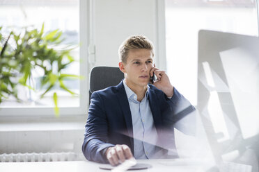 Young businessman sitting at desk in office talking on cell phone - MOEF02159