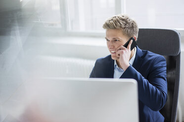 Lächelnder junger Geschäftsmann sitzt am Schreibtisch im Büro und spricht mit seinem Handy - MOEF02160