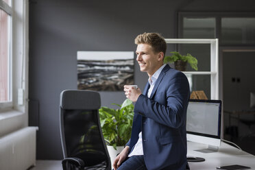 Lächelnder junger Geschäftsmann sitzt auf einem Schreibtisch im Büro und macht eine Kaffeepause - MOEF02167
