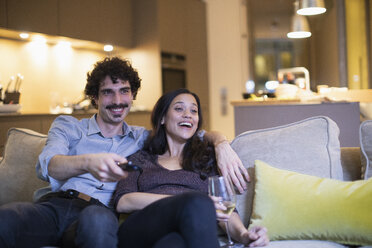 Happy couple watching TV and drinking white wine on living room sofa - CAIF22650