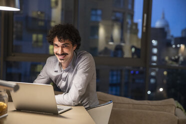 Portrait smiling man using laptop in urban apartment at night - CAIF22654