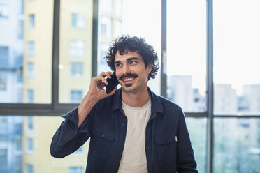 Smiling man talking on smart phone at urban apartment window - CAIF22667