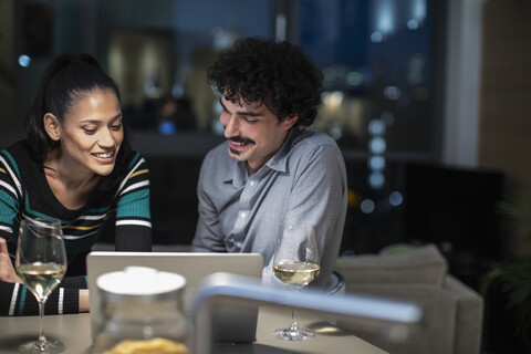 Lächelndes Paar, das abends zu Hause einen Laptop benutzt und Weißwein trinkt, lizenzfreies Stockfoto