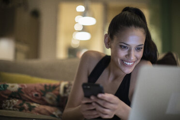 Smiling woman in pajamas using smart phone and laptop on sofa - CAIF22685