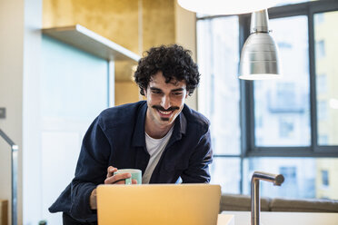 Lächelnder Mann trinkt Kaffee und arbeitet am Laptop in der Wohnküche - CAIF22695