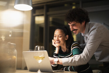 Glückliches Paar, das abends zu Hause einen Laptop benutzt und Weißwein trinkt - CAIF22696