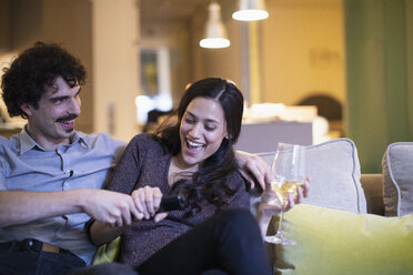 Verspieltes Paar streitet sich um die Fernbedienung, sieht fern und trinkt Weißwein auf dem Sofa - CAIF22698