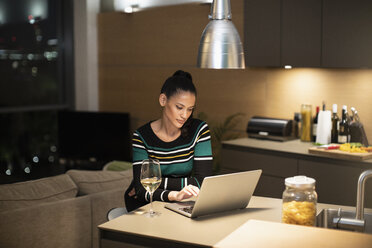 Konzentrierte Frau, die einen Laptop benutzt und Weißwein in der nächtlichen Wohnküche trinkt - CAIF22699