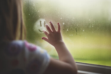 Cropped image of girl making anthropomorphic smiley face on wet window at home during rainy season - CAVF61067