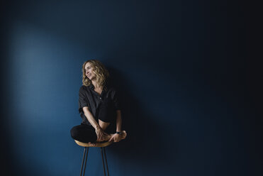 Portrait carefree woman sitting on stool against blue background - CAIF22779