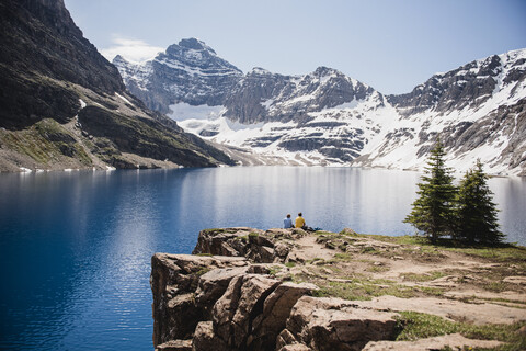 Paar sitzt auf einer Klippe mit Blick auf ruhige, sonnige Berge und einen See, Yoho Park, British Columbia, Kanada, lizenzfreies Stockfoto