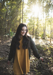 Happy teenage girl looking down while walking against trees in forest - CAVF61075