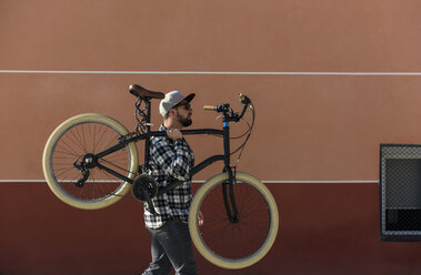 Seitenansicht eines Mannes, der ein Fahrrad trägt, während er an einem sonnigen Tag gegen ein Gebäude in der Stadt läuft - CAVF61084