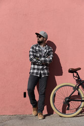 Confident man with arms crossed standing by bicycle on sidewalk against wall during sunny day - CAVF61086